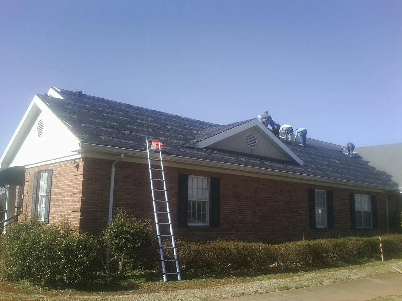 Crew tearing shingles off a brick ranch roof with a ladder set — Emergency Roof Repair work by our crew in Fairlee