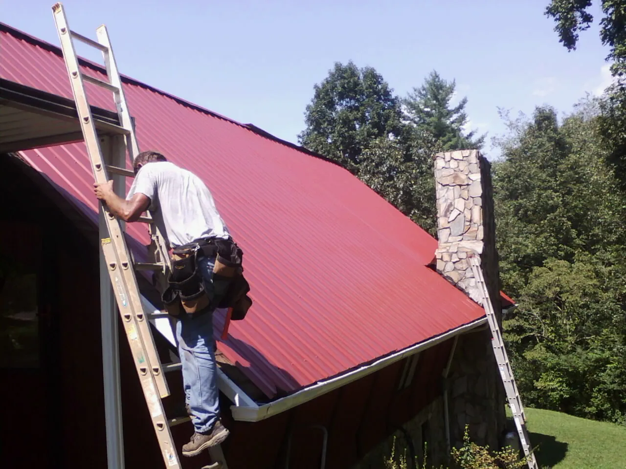 Roofer on a ladder beside a red metal roof and stone chimney — Soffit & Fascia Repair by Roofing Brothers in Fairlee